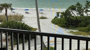 Une vue sur la plage depuis un balcon doté d’une balustrade, avec des palmiers et des parasols de plage.