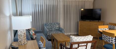 Une chambre d’hôtel avec une table à manger en bois, un canapé, un fauteuil à motifs, une télévision et un ventilateur de plafond.