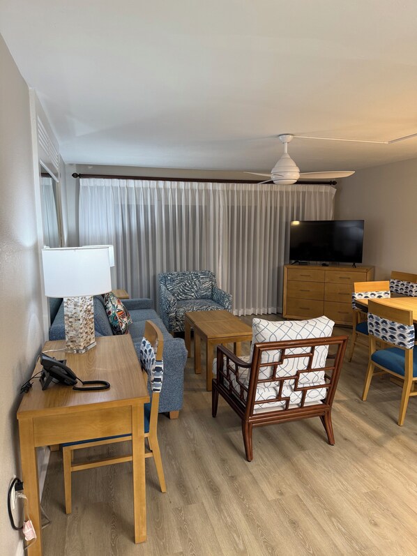 Ein Hotelzimmer mit einem Holztisch, einer Couch, einem gemusterten Sessel, einem Fernseher und einem Deckenventilator.