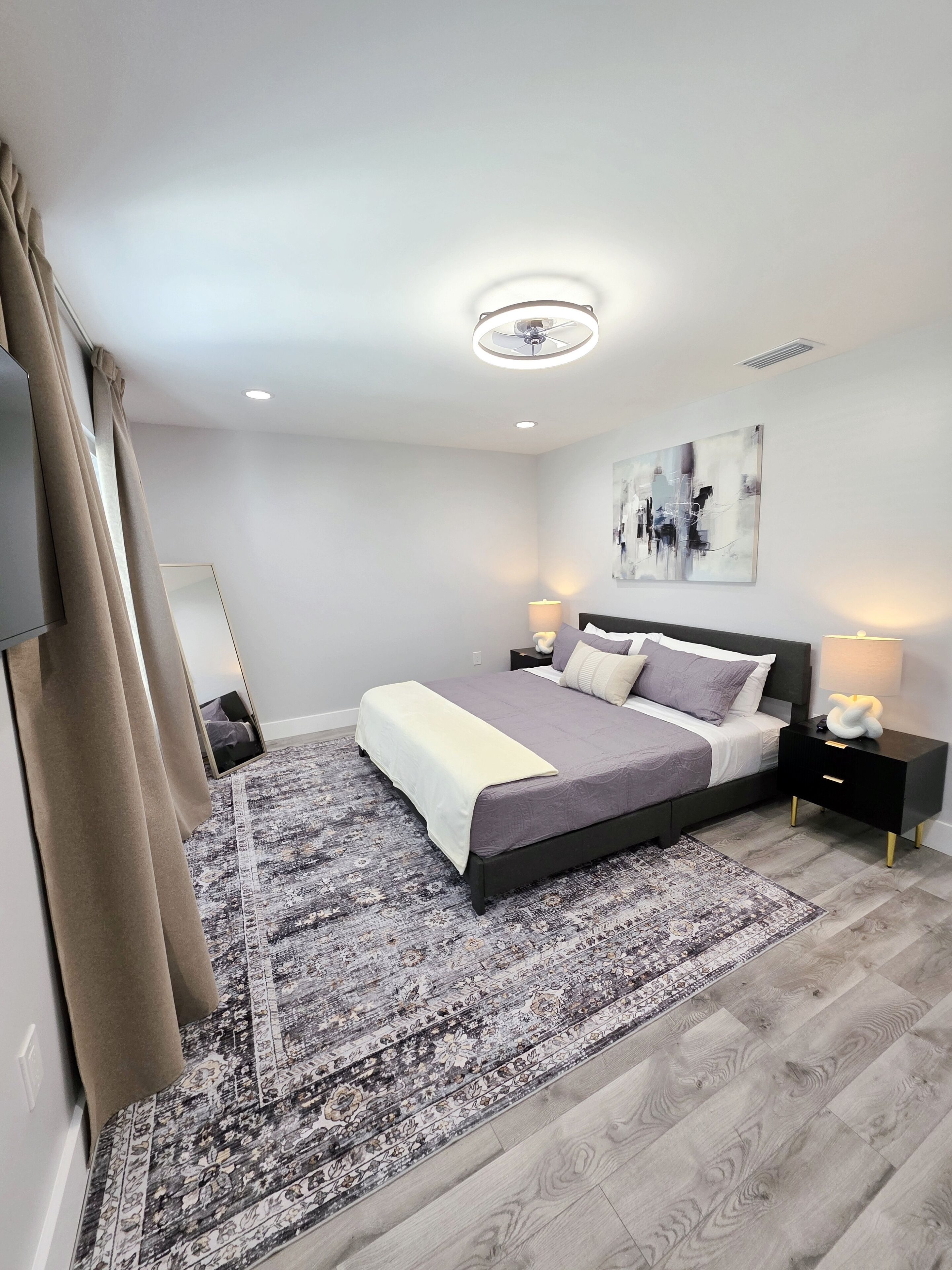 Ein modernes Schlafzimmer mit einem großen Bett, einem gemusterten Teppich, einem Deckenventilator und einem an der Wand befestigten, abstrakten Kunstwerk.