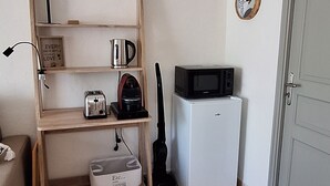 A compact kitchen with a microwave, toaster, kettle, and a small refrigerator. A wooden shelf unit holds kitchenware and a framed quote. A brown woven rug is on the floor, and a door is visible to the right.