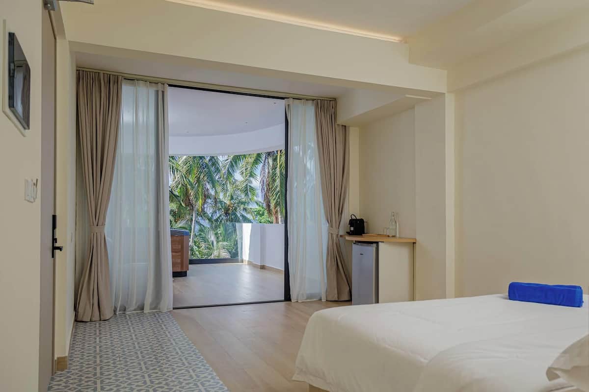 Habitación de hotel con cama, un pequeño escritorio con lámpara, vista a palmeras y puerta corredera que da a un balcón.