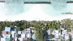 Vista aérea de una zona costera con playa, aguas claras y un conjunto de edificios con techos de varios colores.