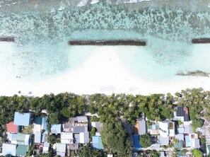 An aerial view of a coastal area with a sandy beach, clear turquoise waters, and a dense cluster of buildings with varied roof colors. - Kinan Boutique (Fulidhoo)