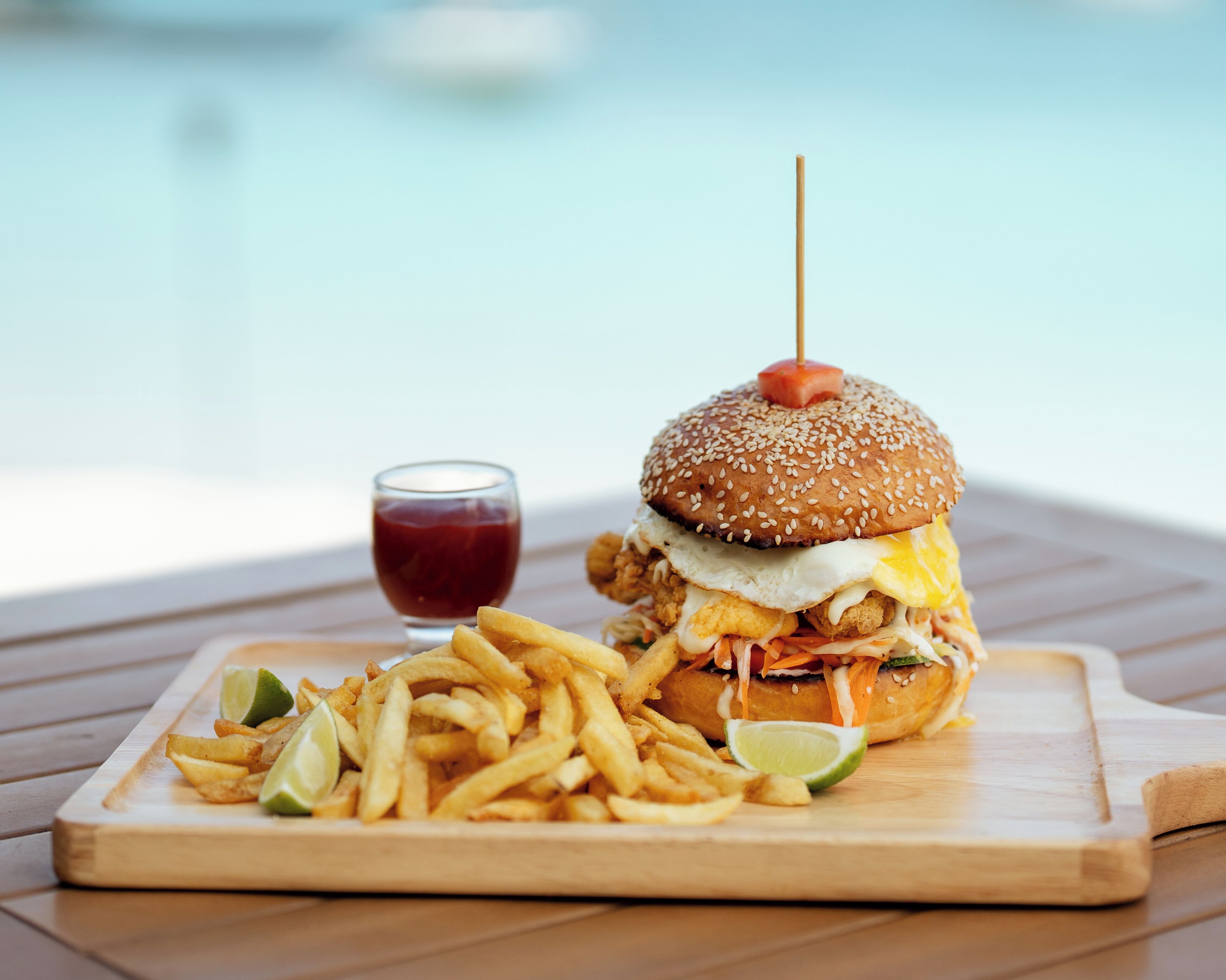 Eine Mahlzeit bestehend aus einem Burger mit Sesambrötchen, einem Spiegelei, Hähnchen und einer Tomate am Spieß, dazu Pommes und ein Glas Ketchup.