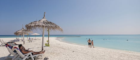 Una playa con arena blanca, agua cristalina y algunas personas. Hay sillas de playa y sombrillas.
