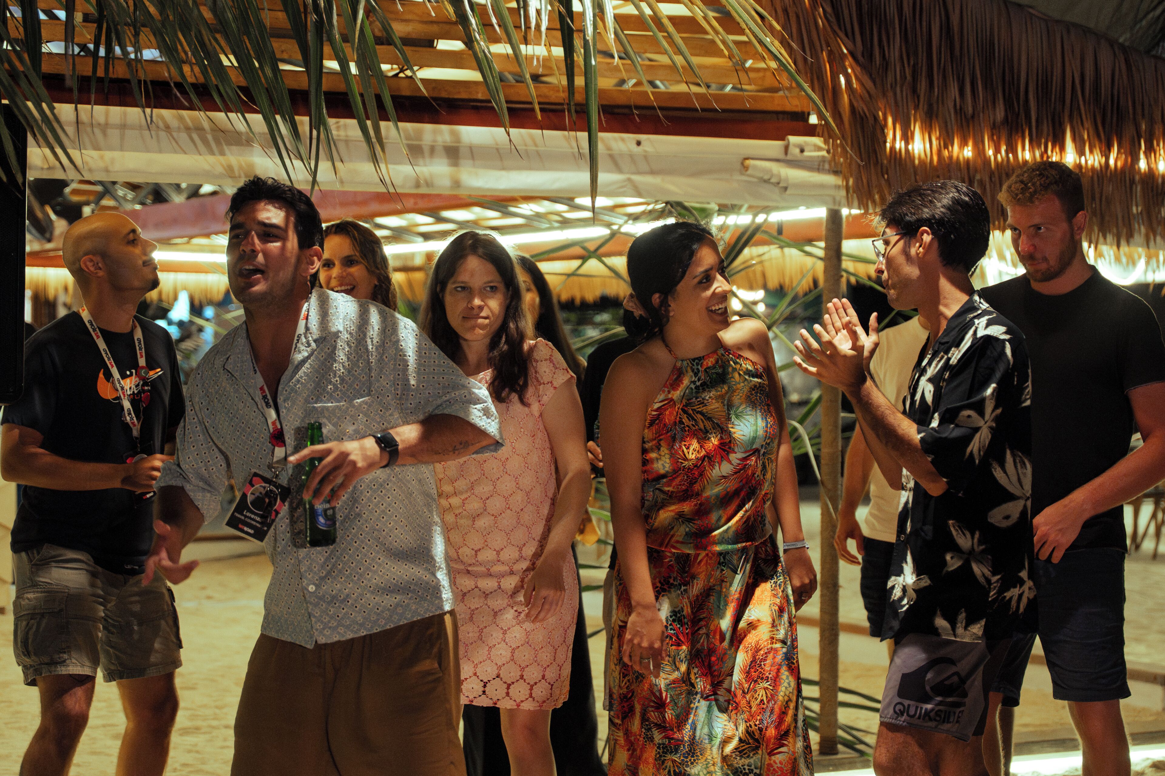A group of people in a covered outdoor area with a thatched roof and palm trees.