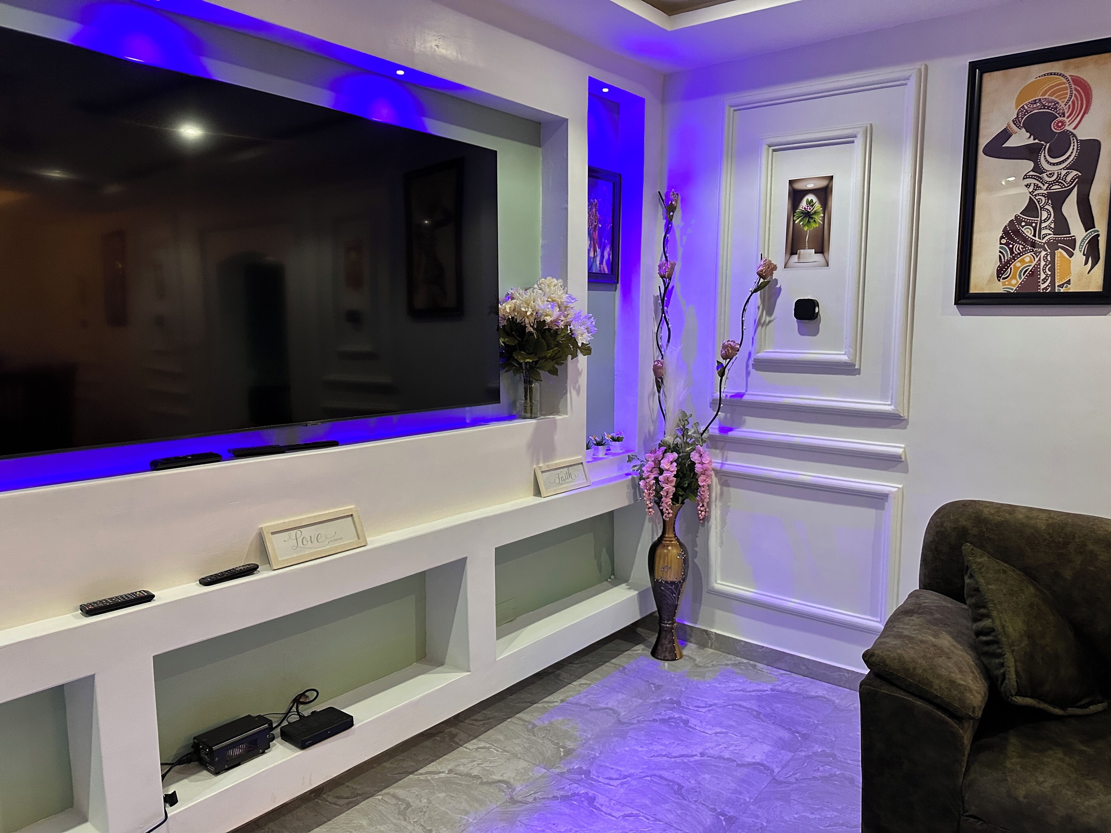 Uma sala de estar com uma televisão de tela plana grande, uma mesa de consola com objetos decorativos, um sofá escuro e uma estante com um vaso de flores.