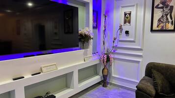 Uma sala de estar com uma televisão de tela plana grande, uma mesa de consola com objetos decorativos, um sofá escuro e uma estante com um vaso de flores.