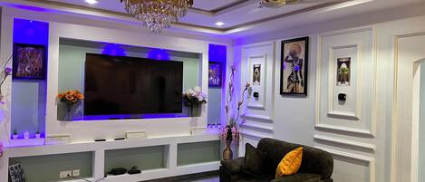 A modern living room with a large flat-screen TV, a dark sofa with a yellow pillow, a black coffee table, and a ceiling fan.