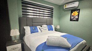 A bedroom with a bed, nightstands, a lamp, an air conditioner, and a framed picture on the wall. - Arek Condos- Your Charming Tropical Home Away from Home in the Heart of Accra. (Greater Accra Region)