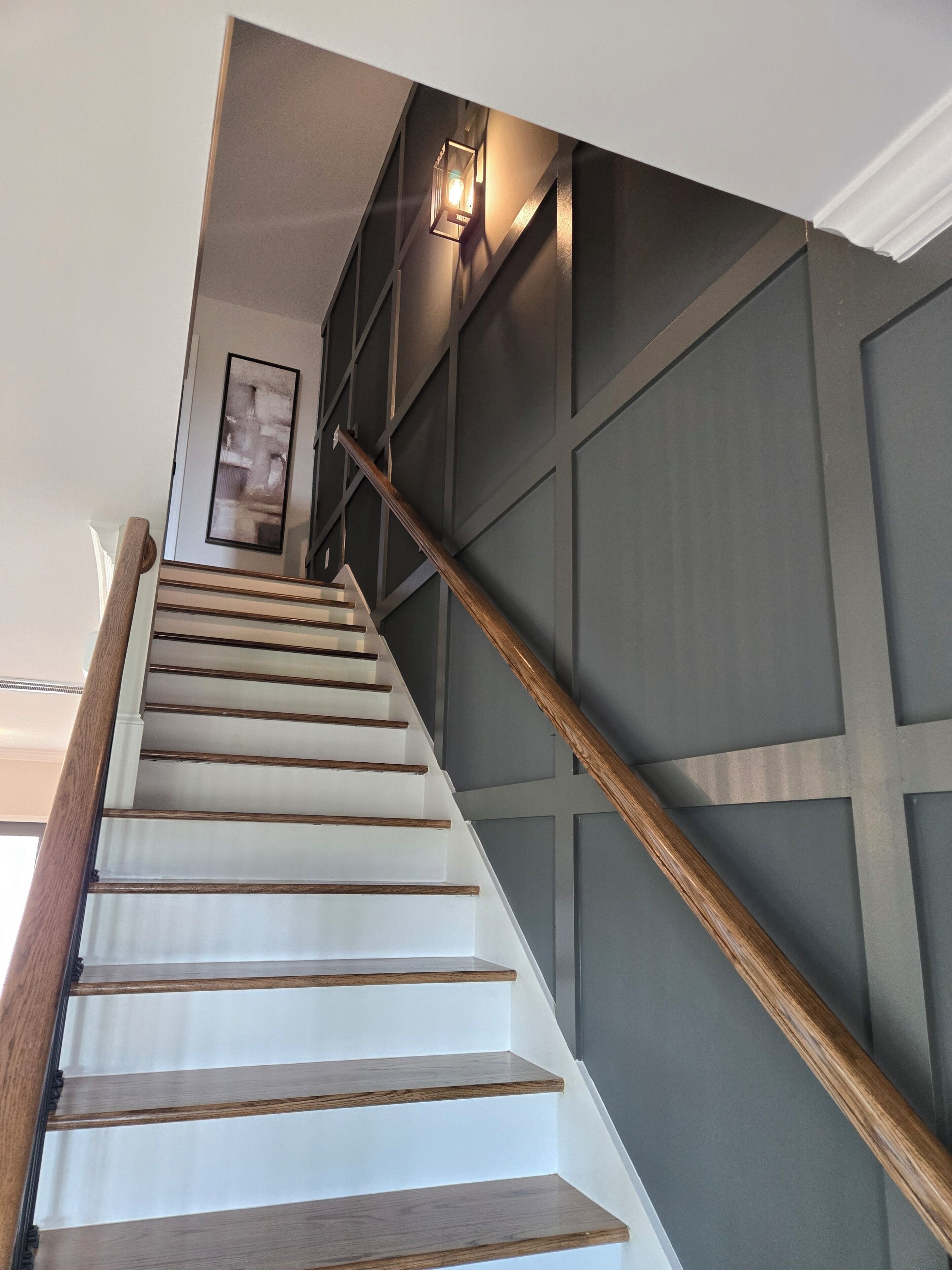 Un escalier avec des marches en bois et une rampe, menant à un palier où est accrochée une image encadrée.