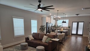 A spacious living room with a brown leather sofa, grey ottomans, and a patterned rug. A dining area with a table and chairs is visible in the background.