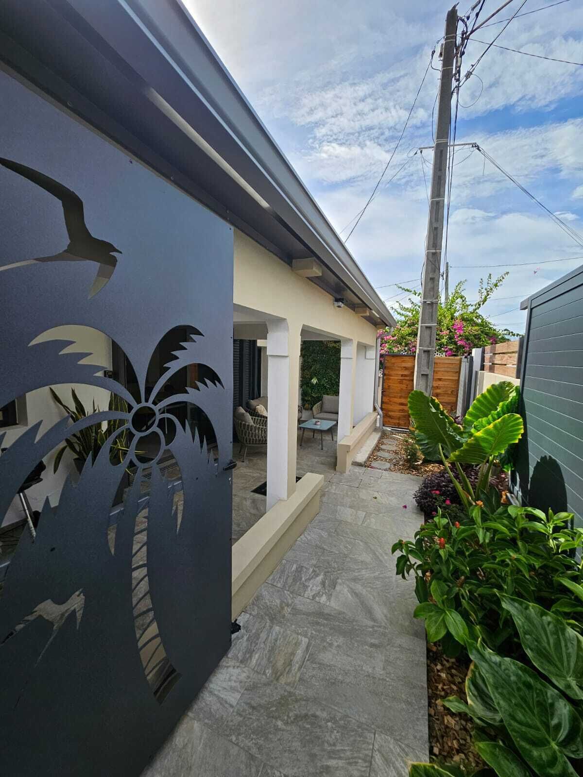 Une maison moderne dotée d’un patio couvert, d’un mur décoratif en métal et d’un jardin planté de grandes plantes à feuilles larges.