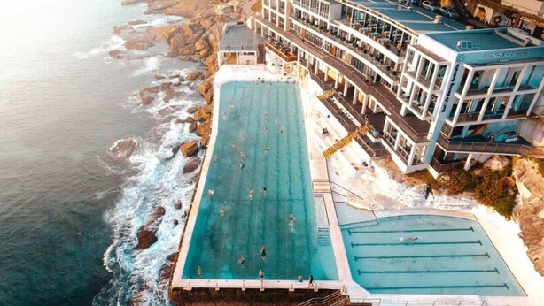 Ein großer, rechteckiger Swimmingpool neben einer felsigen Küstenlinie, mit einem mehrstöckigen Gebäude, das Balkone und Außenbereiche zum Sitzen bietet.