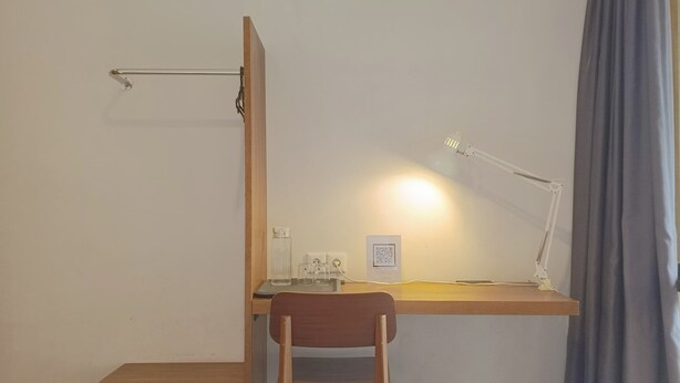 A hotel room with a bed, a desk with a chair, a desk lamp, a small table with a glass, and a QR code on the wall. - Habitat Hills by Santai Aja (Cisarua)