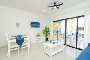 A bright living room with a dining area, a ceiling fan, a TV, and a large window with a view. - Tranquil 2BR Sunshine Bayahibe · Pool · Beach 5 min · AC (San Rafael del Yuma)