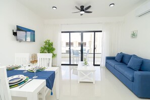 A modern living room with a blue sofa, a dining table set with plates and glasses, a ceiling fan, and a TV mounted on the wall. - Tranquil 2BR Sunshine Bayahibe · Pool · Beach 5 min · AC (San Rafael del Yuma)