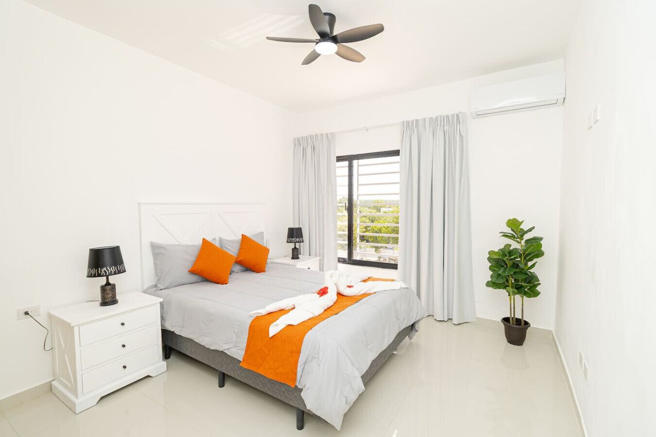 Une chambre à coucher comprenant un lit, des tables de chevet, un ventilateur de plafond, une fenêtre avec des rideaux et une plante en pot.
