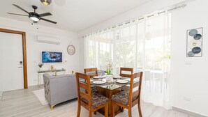 Un comedor con una mesa de vidrio, cuatro sillas de madera, un sofá gris, un ventilador de techo y un televisor en la pared.