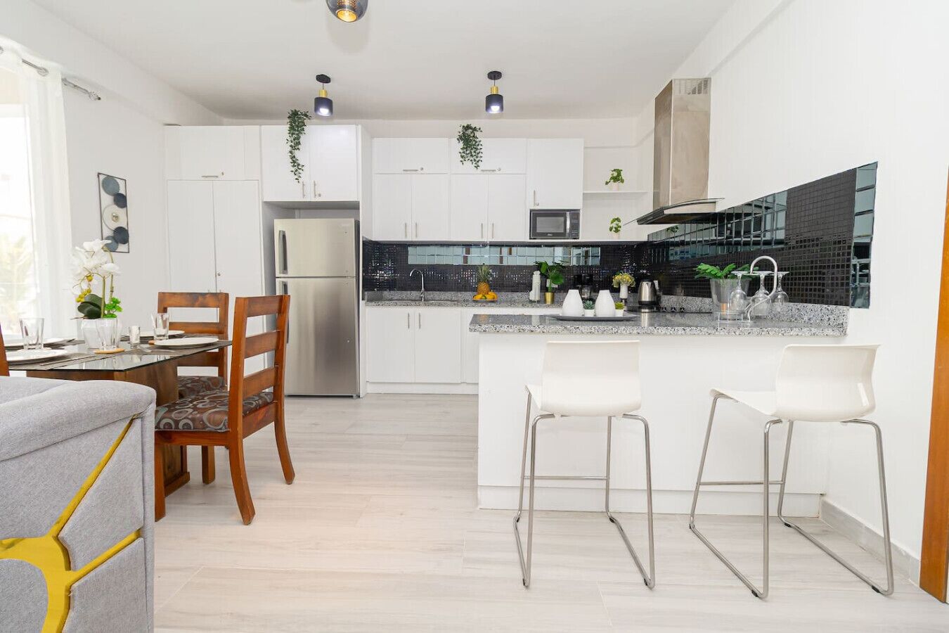 A modern kitchen with white cabinets, a stainless steel refrigerator, and a black backsplash. There are two white bar stools and a wooden dining table with chairs.