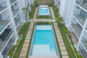 Um hotel moderno com duas piscinas retangulares, rodeado por um gramado bem cuidado e palmeiras.