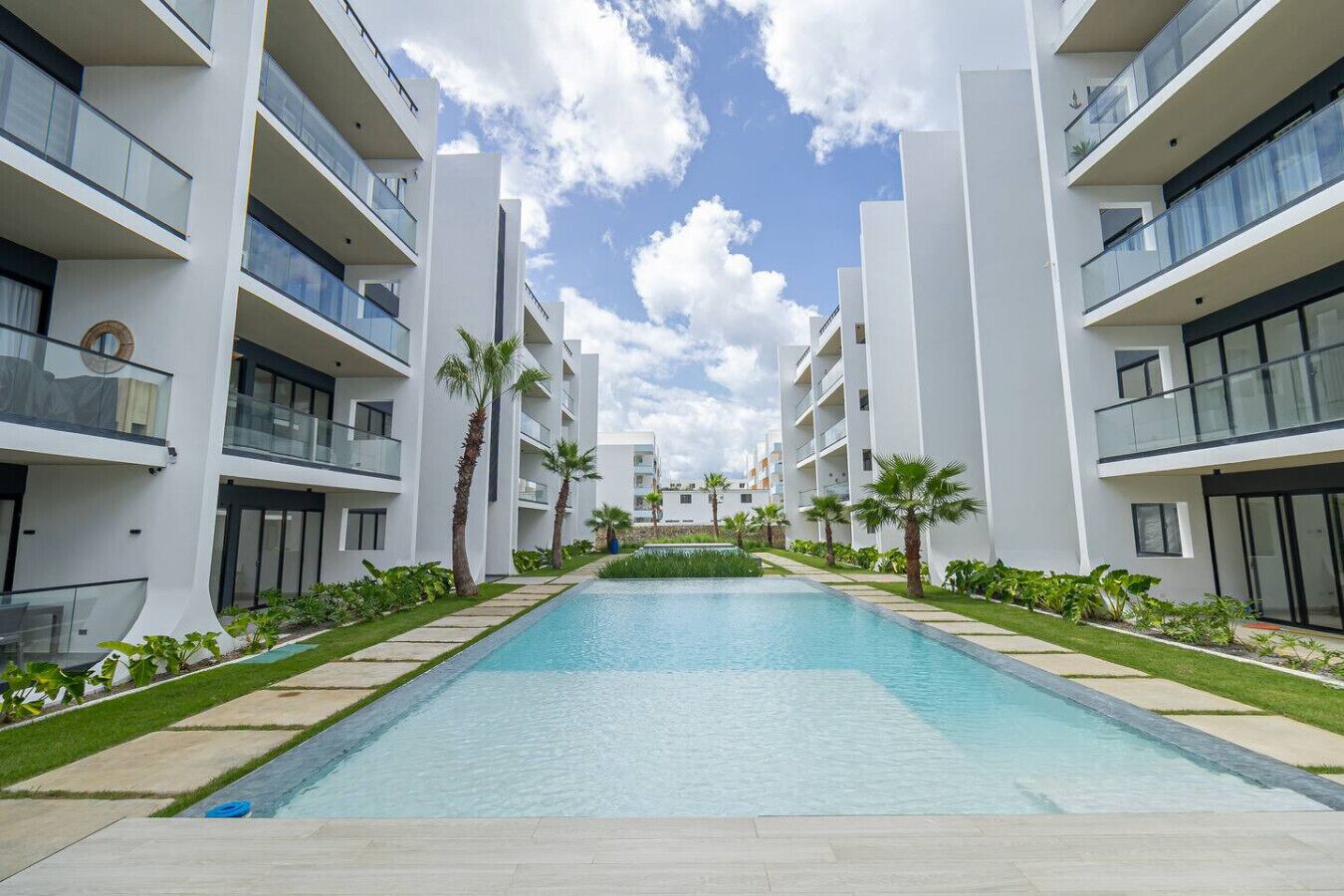 A modern hotel with a central swimming pool, surrounded by palm trees and greenery.