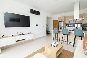 Un salon moderne doté d’un téléviseur à écran plat, d’une table basse en bois et d’un coin cuisine avec des tabourets de bar.