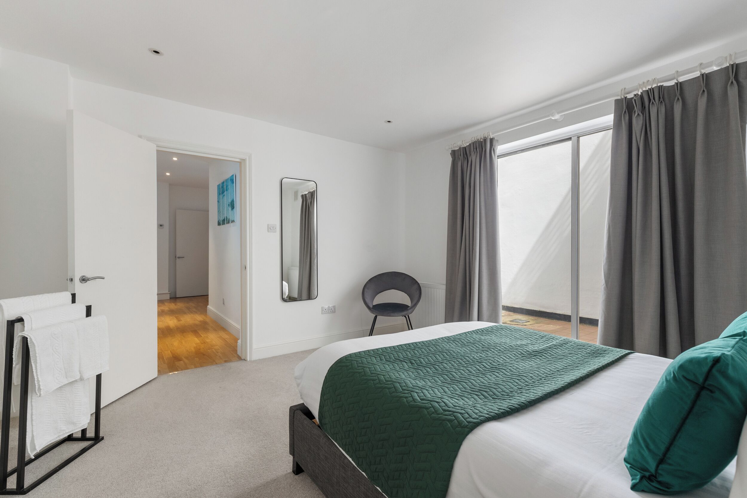 A bedroom with a bed, a bedside table, a chair, a mirror, and a view of the outdoors.