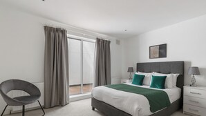 Ein Schlafzimmer mit Bett, Stuhl, Nachttisch, Lampe und einem Fenster mit Vorhängen.