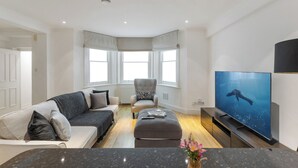 Ein modernes Wohnzimmer mit Sofa, Sessel, Hocker und einem Flachbildfernseher.