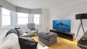 Ein modernes Wohnzimmer mit Sofa, Hocker, Sessel und einem Flachbildfernseher auf Holzdielen.