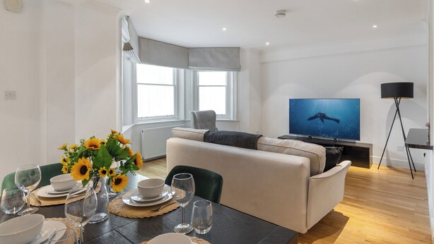 Ein modernes Wohnzimmer mit einer Couch, einem Esstisch für vier Personen, einem Flachbildfernseher und einem großen Fenster mit Vorhängen.