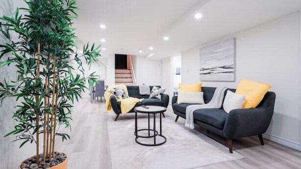 A modern living room with a large potted plant, two sofas with cushions, a small round table, and a staircase leading to another room. - Stylish, Spacious 6-Bedroom Home in the GTA (Whitby)