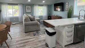 A modern kitchen with a central island, a dining area with wicker chairs, a sofa, and a TV mounted on the wall. - Longfellow Tides (Lexington)