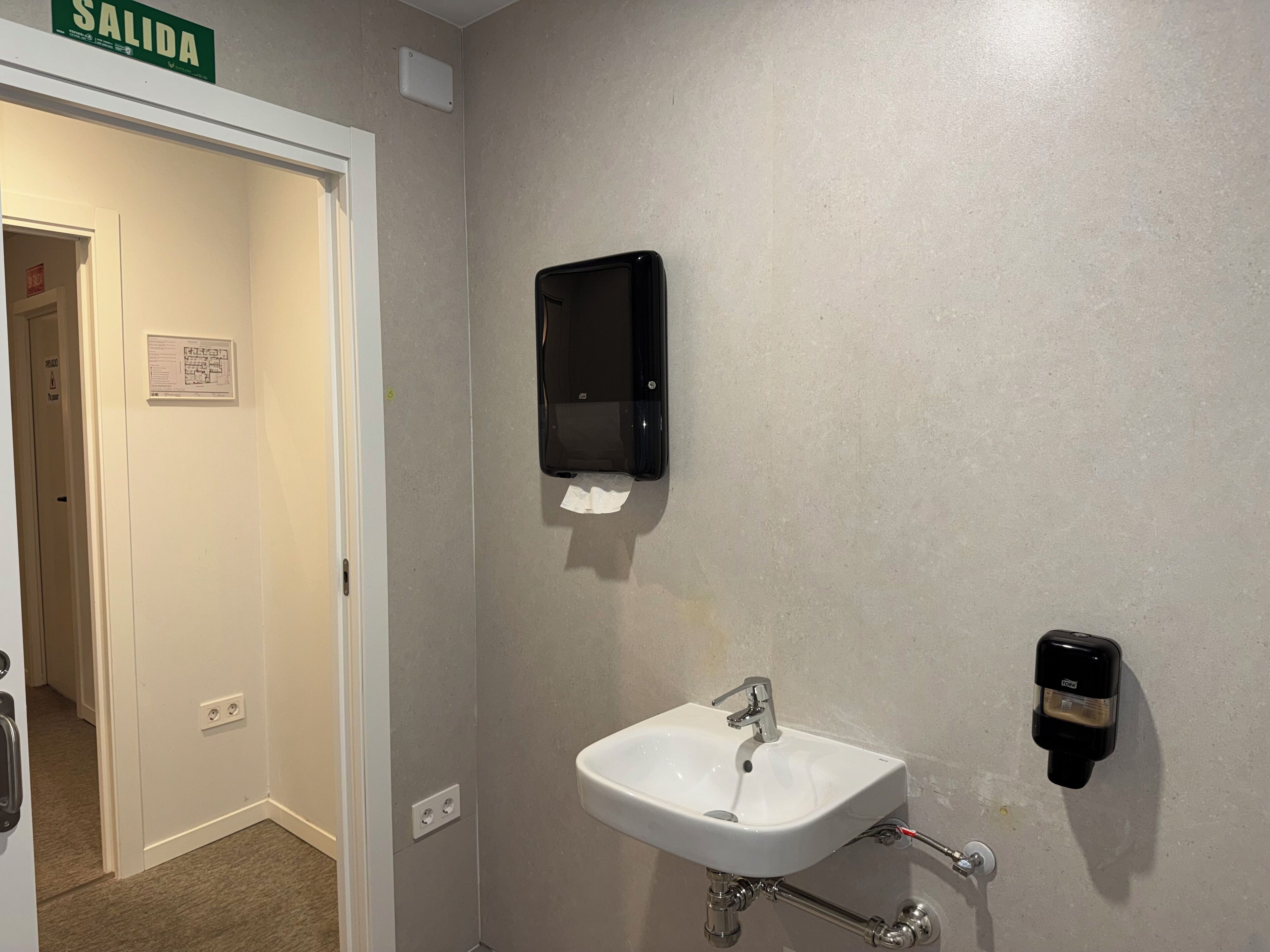 Un baño con un lavamanos blanco, un dispensador de toallas de papel y un dispensador de jabón en la pared.