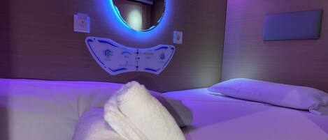 Una habitación con una cama, un espejo circular con iluminación azul y un panel de control en la pared.