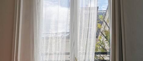A room with a bed, a window with sheer curtains, and a view of the outside.