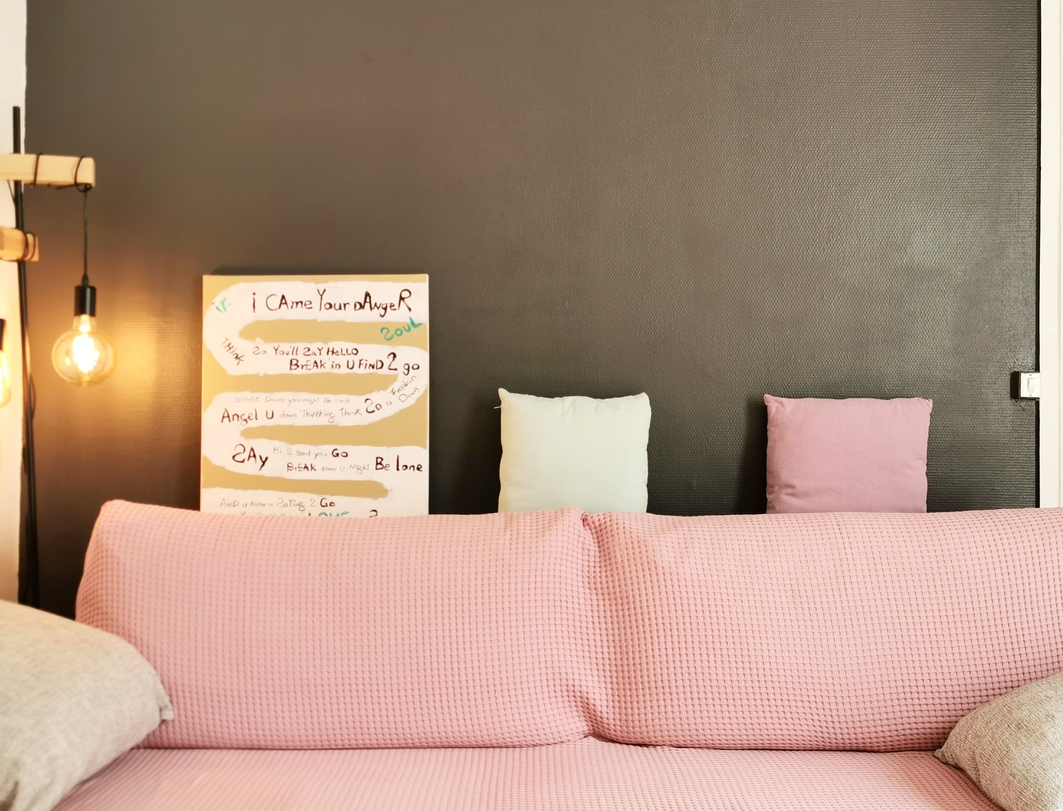 A bedroom with a pink textured sofa, two pillows, a wall-mounted light fixture, and a motivational poster.