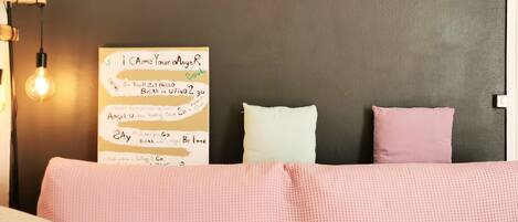 A bedroom with a pink textured sofa, two pillows, a wall-mounted light fixture, and a motivational poster.