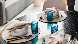 Ein Esstisch ist gedeckt mit blauem Glas, Tellern und Besteck auf gewebten Platzsets, im Hintergrund ein graues Sofa und eine Treppe.
