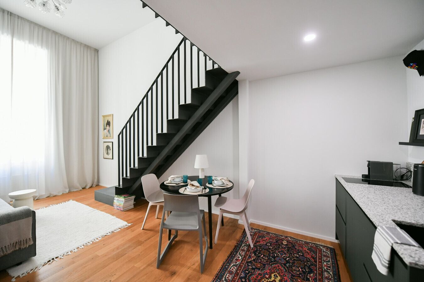 A modern living room with a staircase, a dining area with chairs and a table, and a kitchen area with a countertop and appliances.
