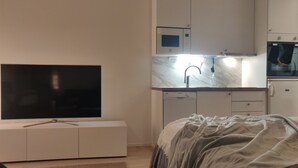 A modern bedroom with a TV on a white cabinet, a bed with a gray blanket, and a kitchen area with a sink and cabinets.