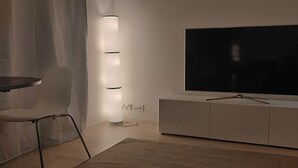 A modern living room with a flat-screen TV mounted on a white cabinet, a standing lamp, a dining table with a chair, and a patterned rug.