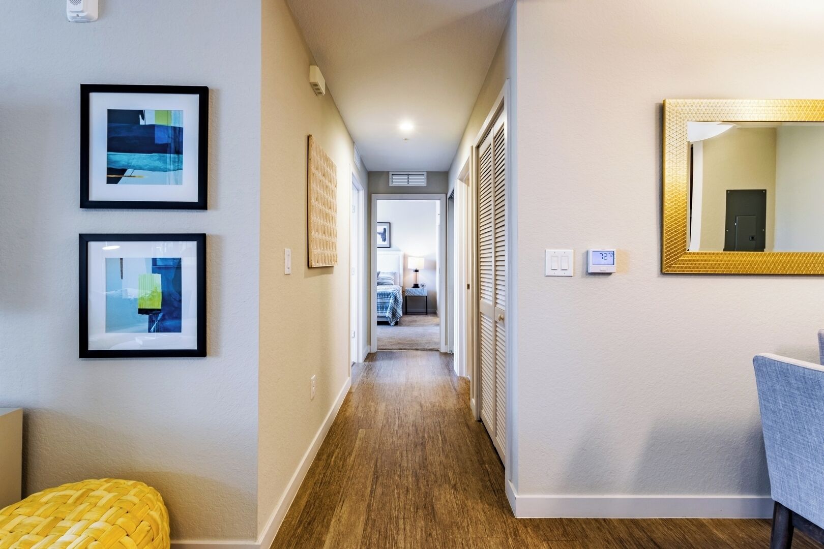A hallway with framed artwork, a yellow woven bench, a mirror with a gold frame, and a door leading to a bedroom.