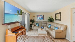A living room with a large TV displaying a beach scene, a beige sofa, a wooden desk, and a small coffee table. - Myrtle Beach Resort Ocean Spa 351- 2BR-Pool (Myrtle Beach)