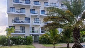 A modern multi-story building with balconies, a paved walkway, and palm trees.
