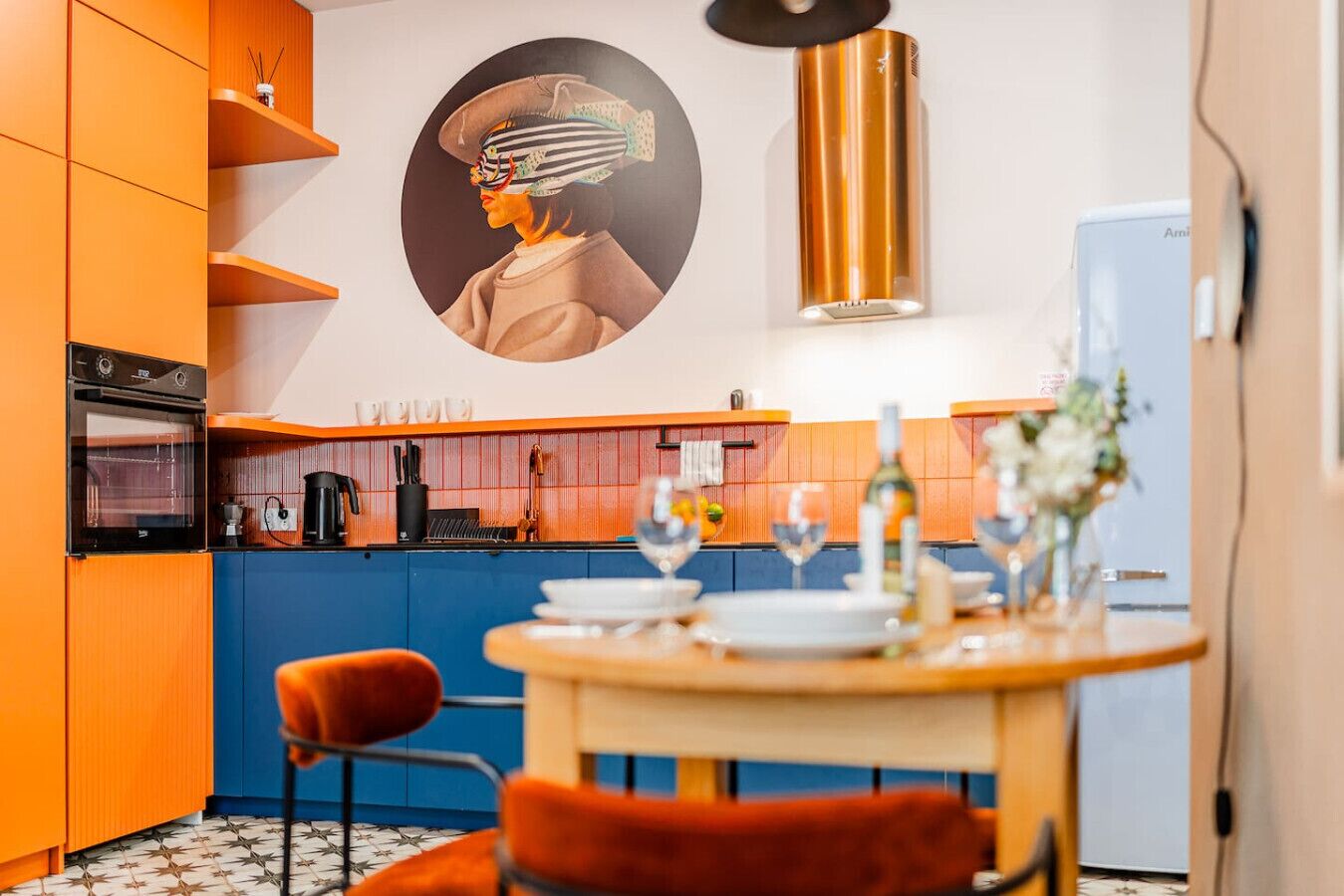 A modern kitchen with orange cabinetry, a built-in oven, and a dining area with a table set for two.