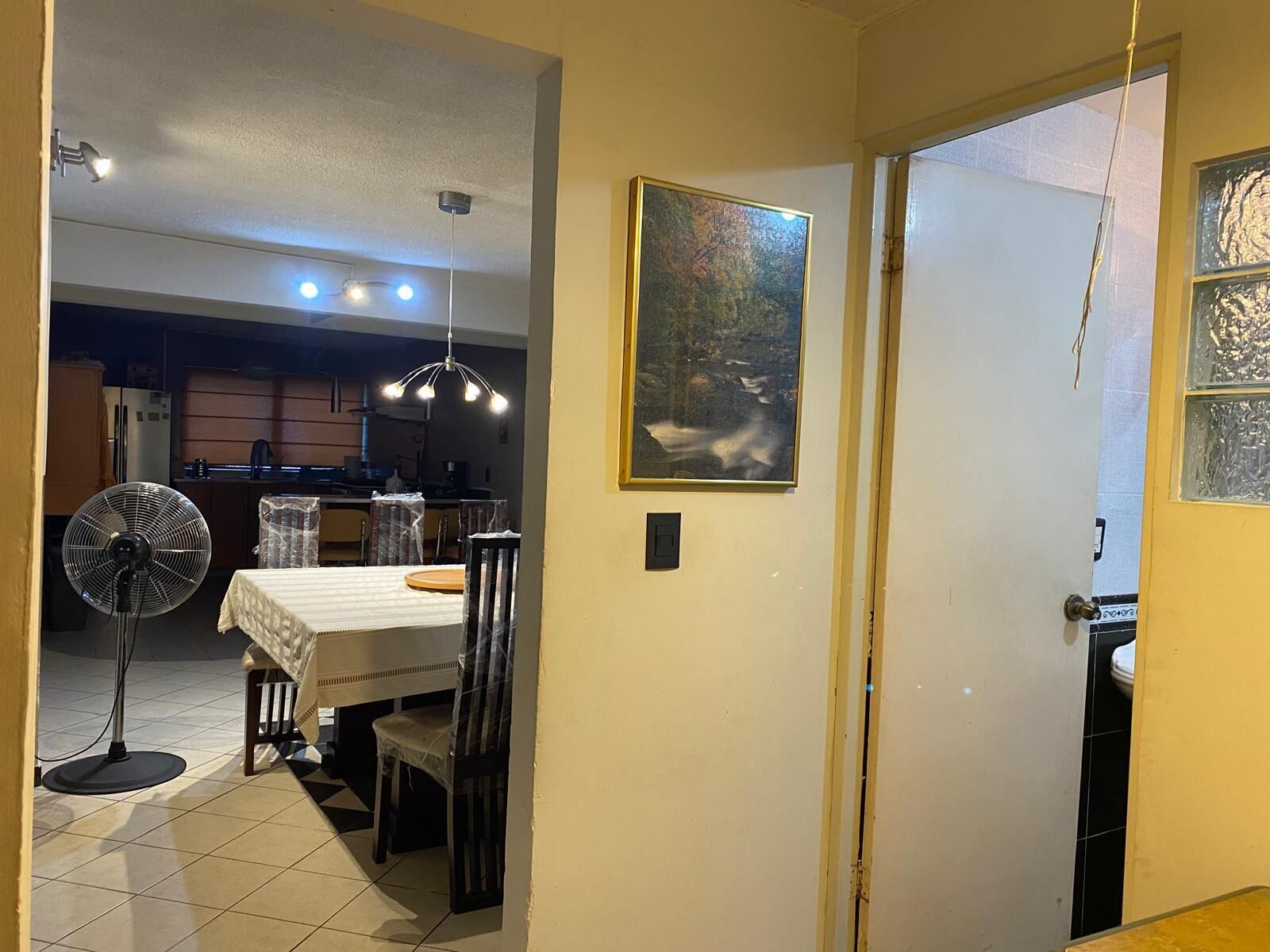 A dining area with a table, chairs, a fan, and a kitchen in the background.