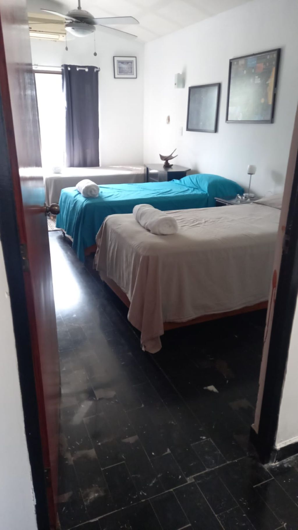 A room with two beds, one with a blue bedspread and the other with a beige bedspread. There is a ceiling fan, a window with curtains, and a framed picture on the wall.